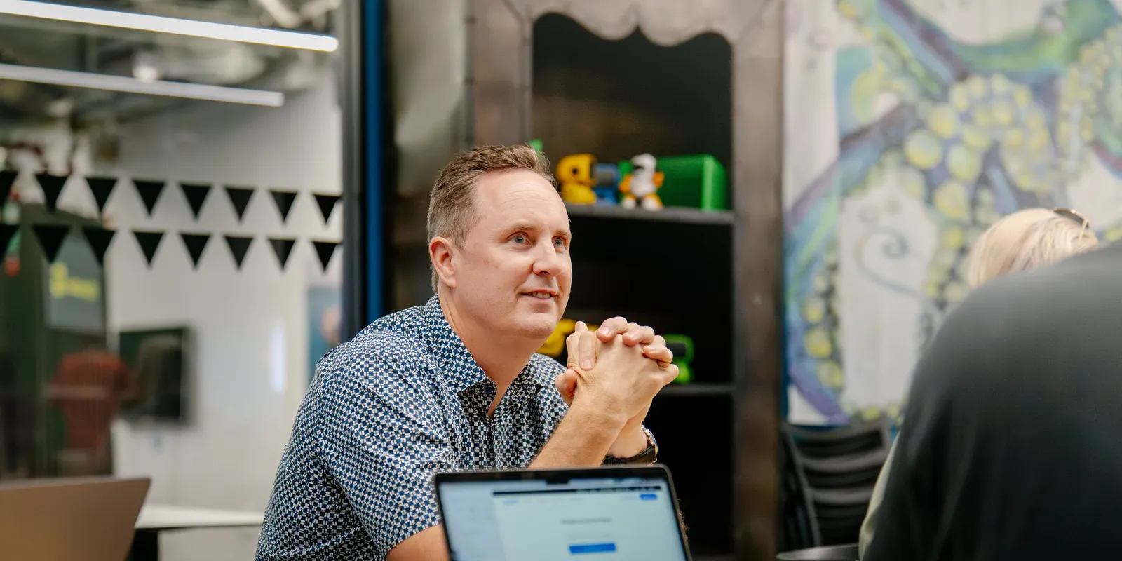 John Halvorsen-Jones listening across a meeting table, hands clasped, a laptop open in front of him
